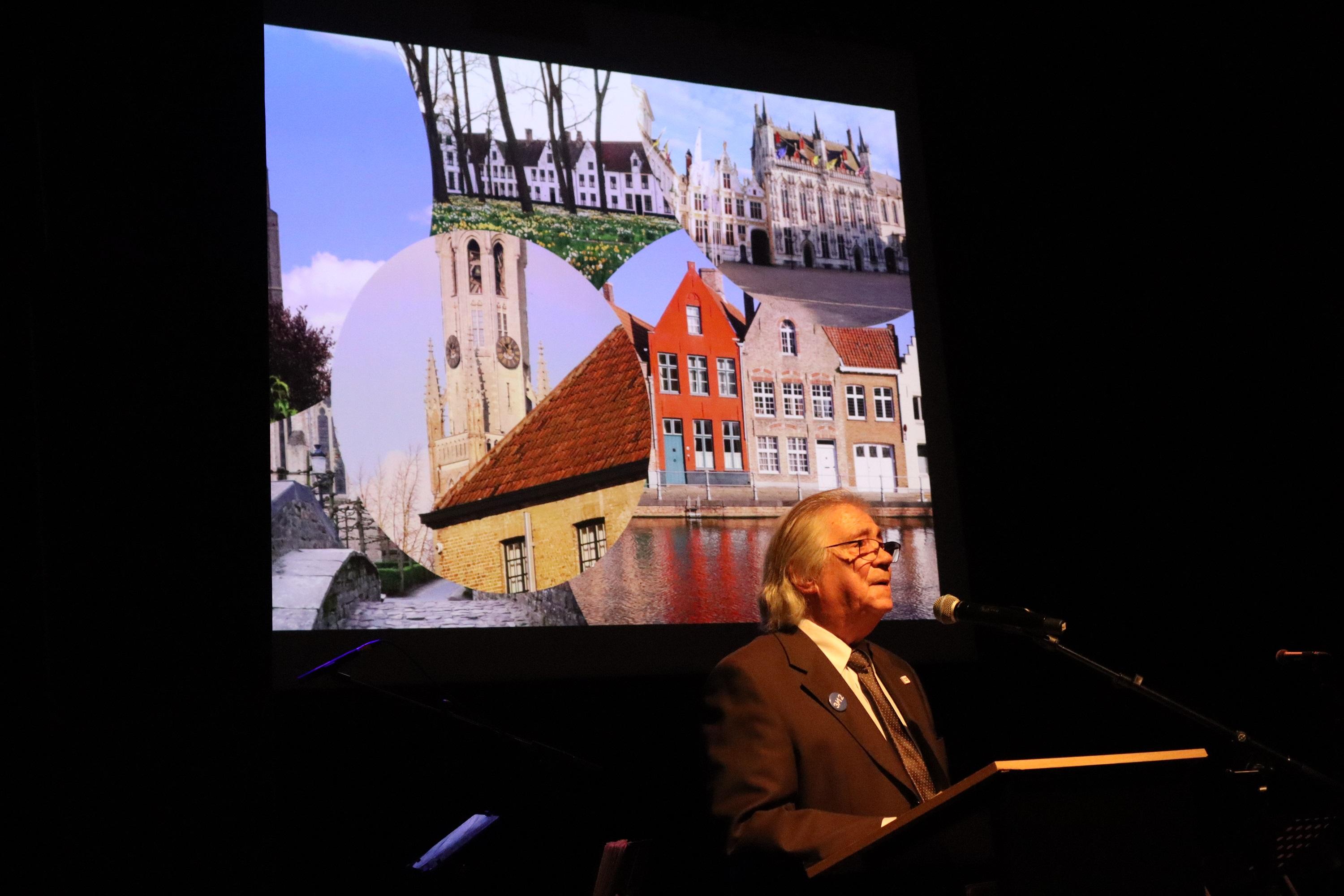 Een wat oudere man staat achter het spreekgestoelte op het podium. Achter hem worden verschillende foto\'s van de stad Brugge geprojecteerd.