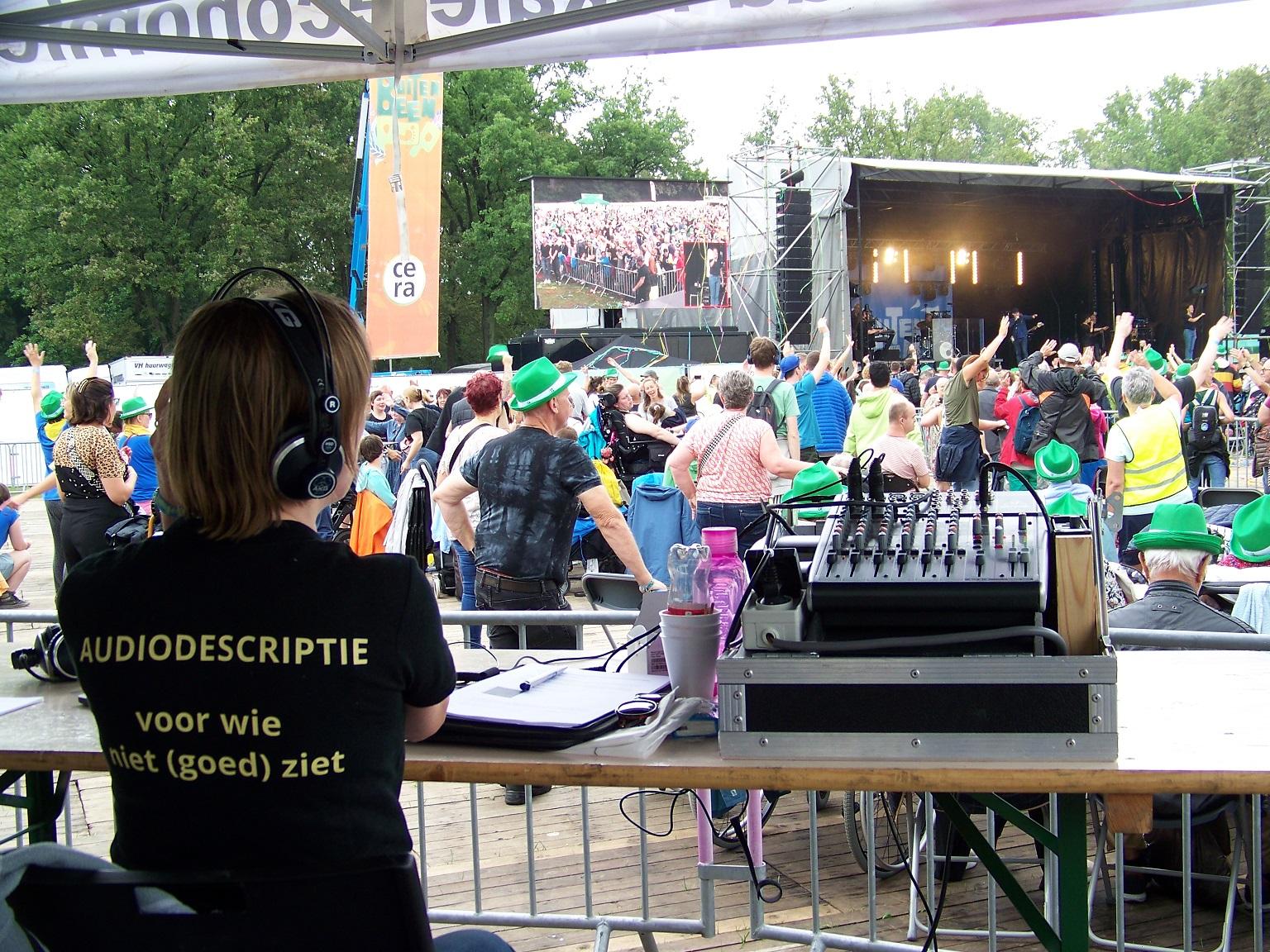 Audiobeschrijver Susanne heeft een headset op en kijkt naar het podium met de artiesten. Achter op haar shirt staat \'audiodescriptie voor wie niet (goed) ziet\'.