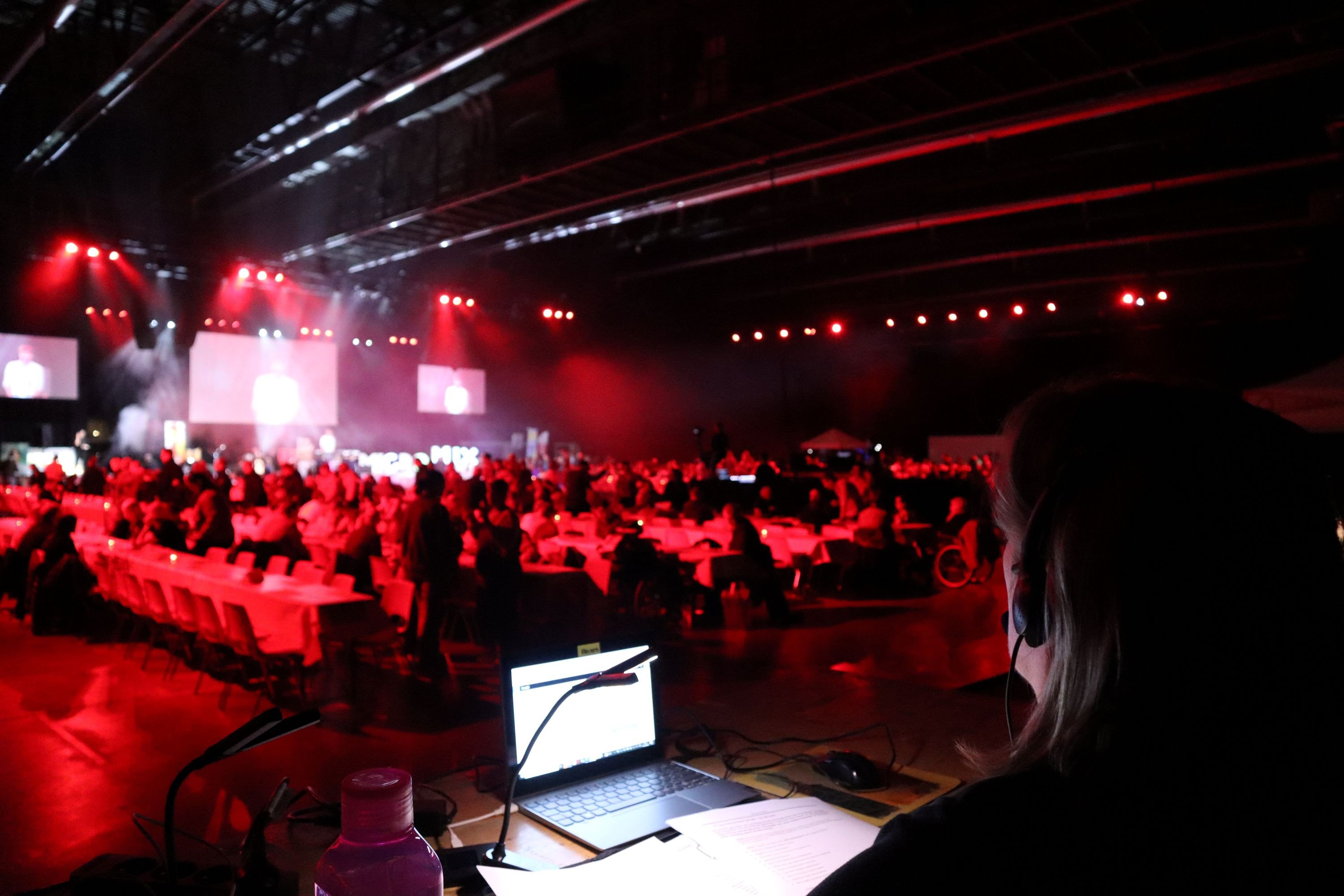 Susanne zit aan haar bureau met een headset op en het script voor haar, net zoals haar laptop. Op de achtergrond het Micromix-podium waarop de artiesten optreden met daarvoor een kleine dansvloer en vele rijen tafels waar de gasten aan zitten. De zaal baadt in rood licht.