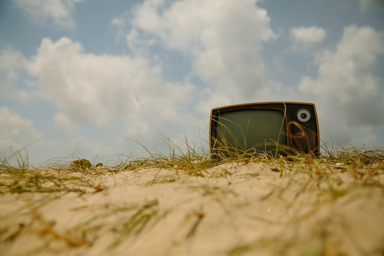 Een ouderwetse tv ligt in een duinlandschap met op de achtergrond enkele wolkenplukjes.