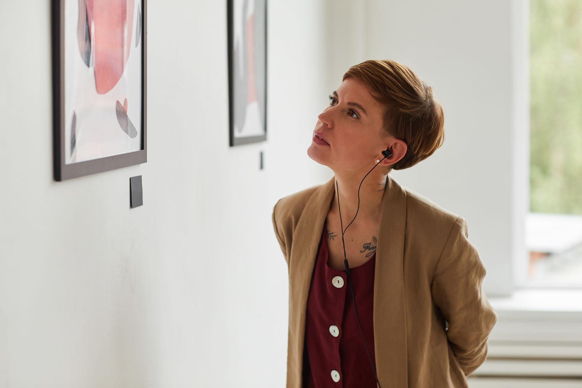 Een jonge vrouw in een museum luistert naar een audiogids terwijl ze een schilderij bestudeert. Ze heeft kort bruin haar en tatoeages in haar nek. Ze draagt een donkerrood shirt en een beige blazer.