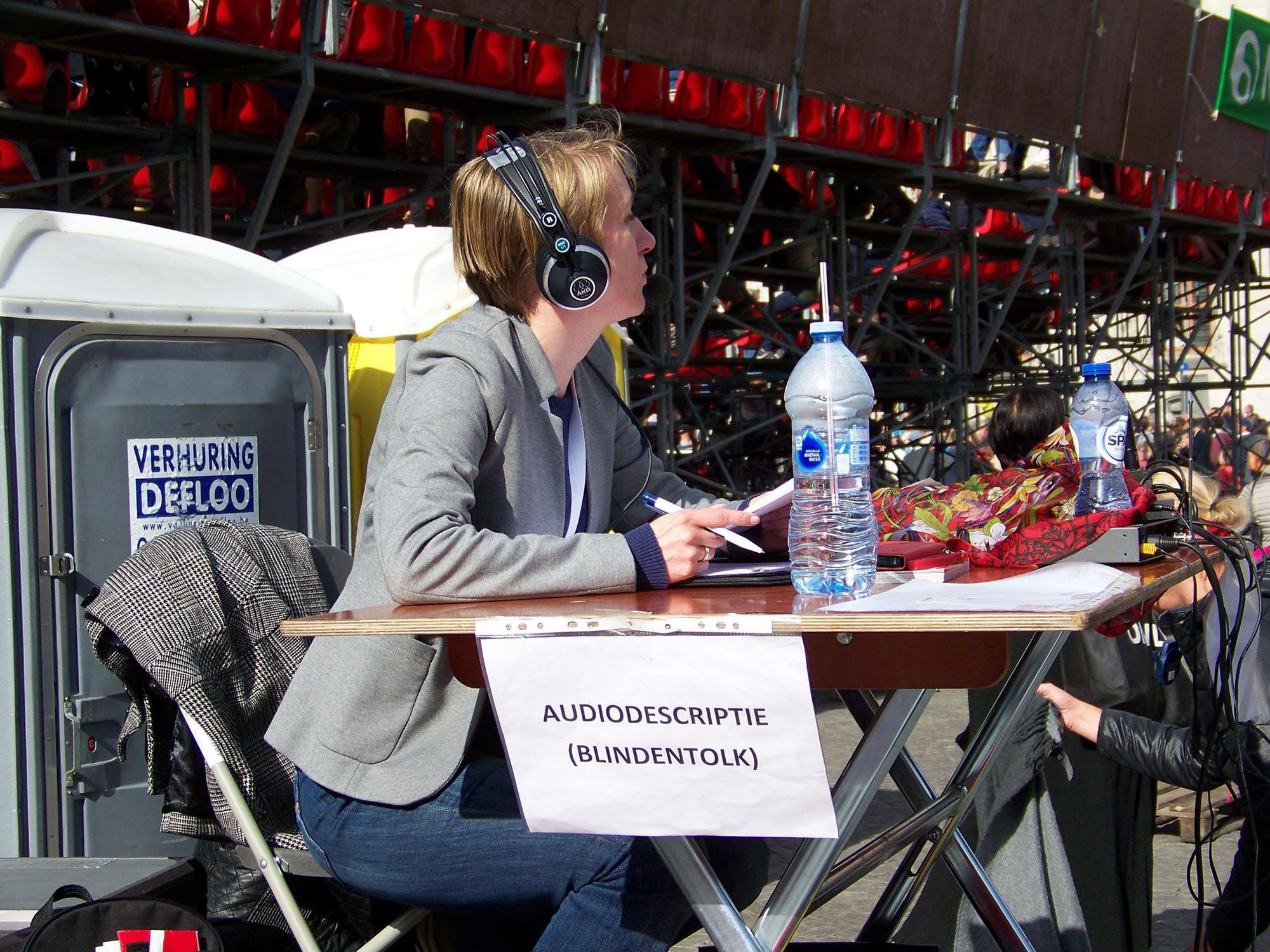 Audiobeschrijver Susanne Verberk heeft een headset op. Aan haar tafel hangt een bordje met de tekst \'audiodescriptie / blindentolk\'.