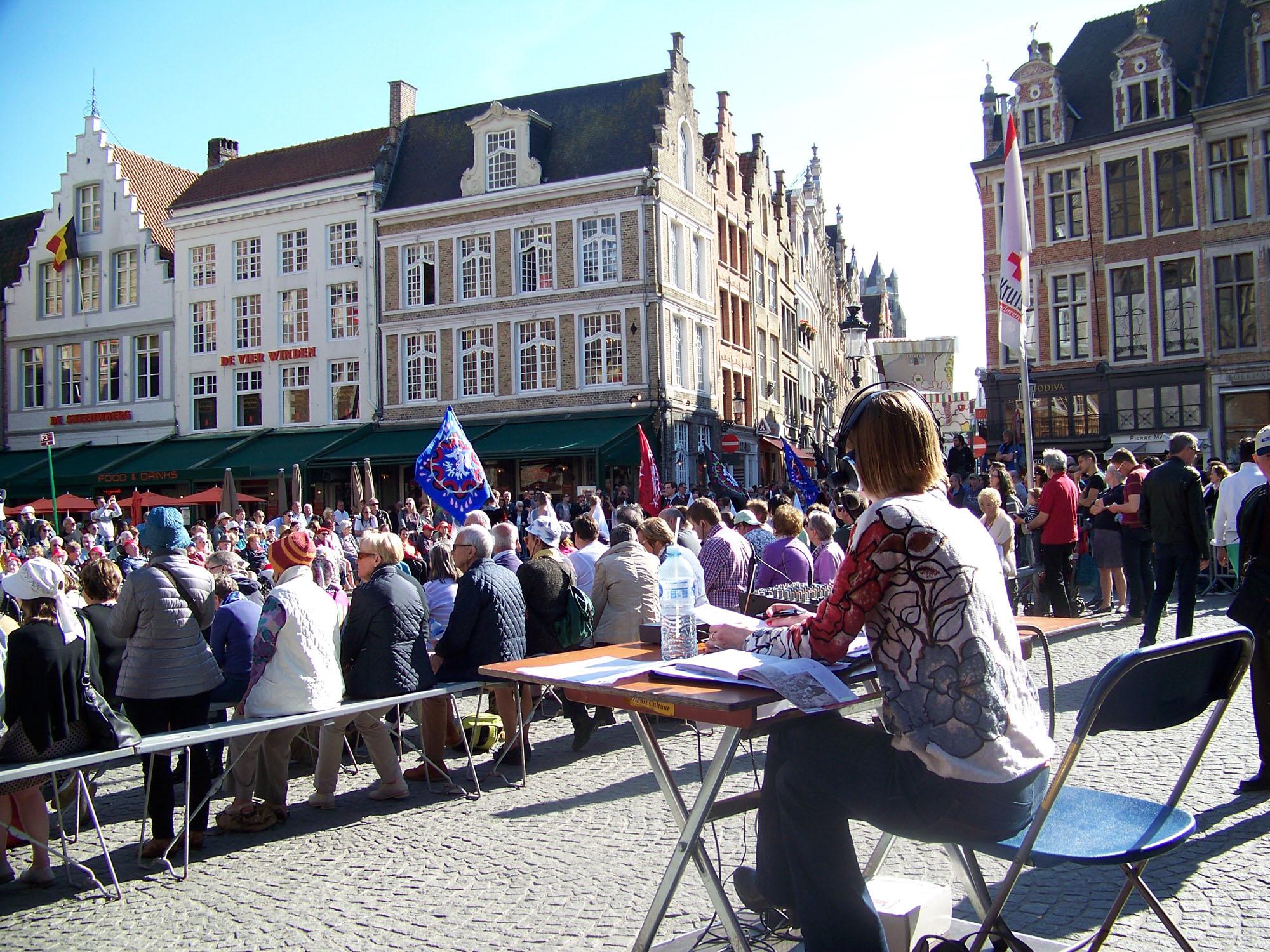 Susanne zit aan een tafel op een klein podium achter het publiek. Op de achtergrond trekt de Heilig Bloedprocessie voorbij.